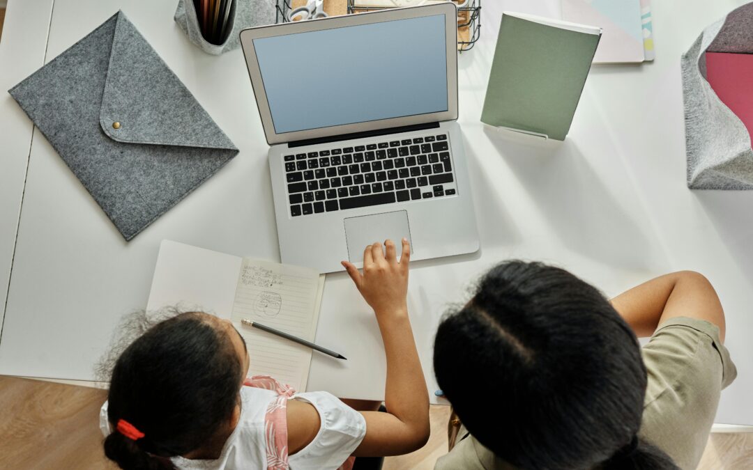 Vue de dessus d’une personne travaillant sur un ordinateur portable avec un enfant, un carnet et des fournitures de bureau posés sur le bureau.