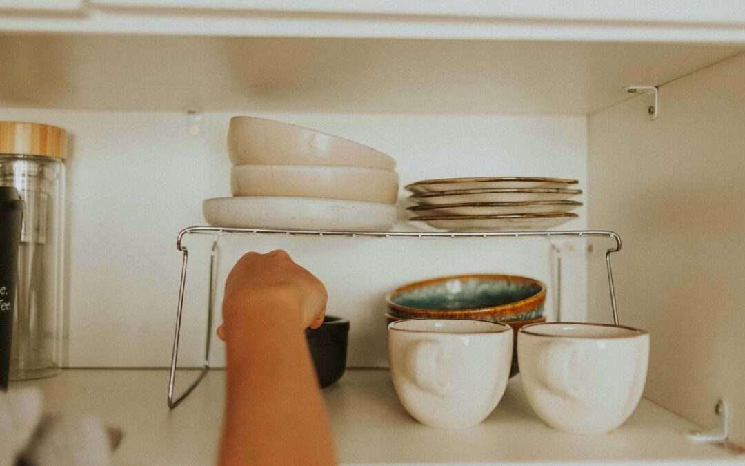 Une personne range de la vaisselle dans une étagère de cuisine, avec des tasses, des bols et des assiettes empilés sur une étagère métallique.
