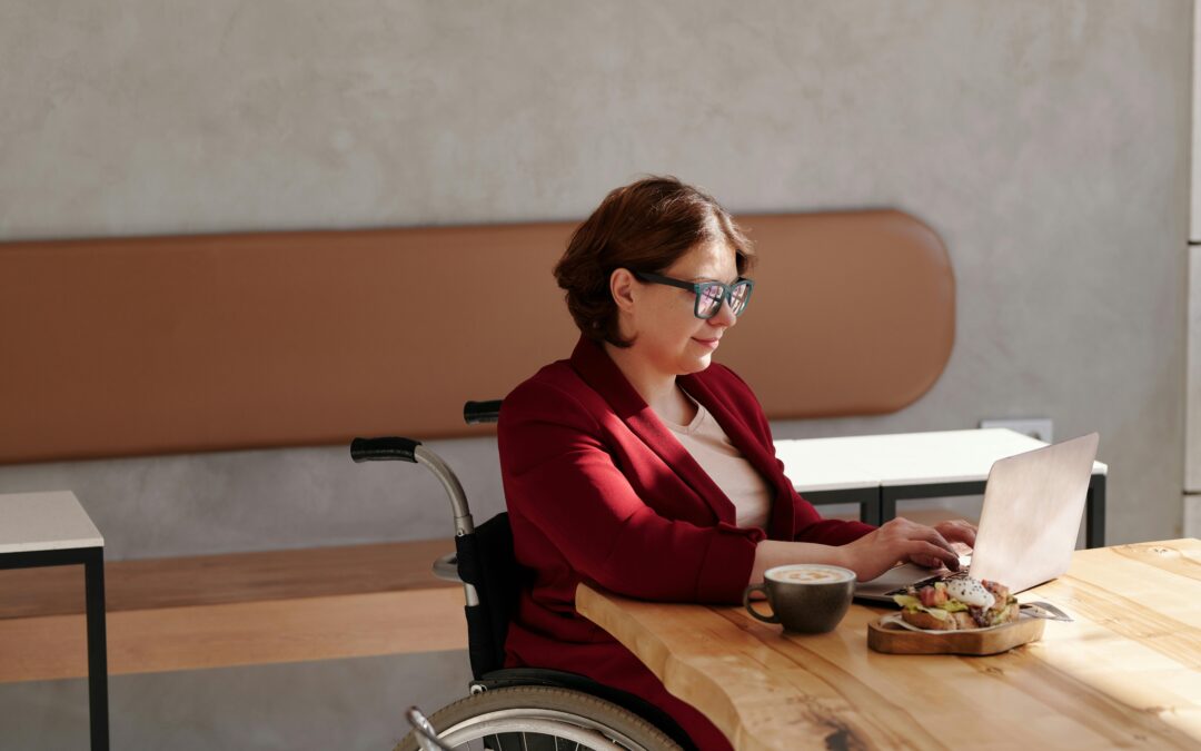 Une femme assise dans un fauteuil roulant travaille sur un ordinateur portable dans un café, vêtue d’une veste rouge. Une tasse de café et une assiette de toast sont posées devant elle sur la table en bois claire